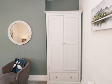A white wardrobe next to a gray chair with a bird cushion and a round mirror on the wall at Derwent Cottage in Keswick