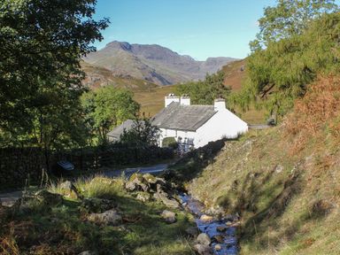 A rural cottage beside a small stream with trees and hills in the background at Derwent Cottage in Keswick
