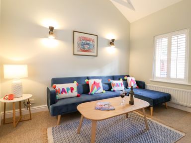 A living room with a blue sofa and colorful cushions a wooden coffee table with wine glasses and a bottle a side table with a lamp and window shutters at Poppy Cottage Southwold