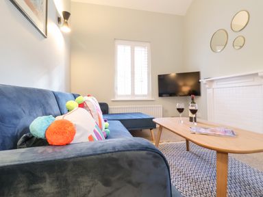 A living room with a blue sofa and colorful pillows a wooden coffee table with two glasses of red wine a bottle and magazines a wall mounted TV and three round mirrors above a white fireplace at Poppy Cottage in Southwold
