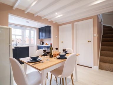 A dining area with a wooden table set for four including plates bowls wine glasses and cutlery next to a kitchen with blue cabinets a sink and a white refrigerator at Poppy Cottage in Southwold