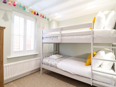 A bedroom with white bunk beds yellow pillows a window with shutters and a colorful tassel garland at Poppy Cottage in Southwold