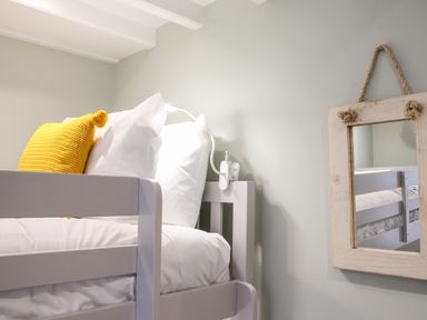 A bedroom with a bunk bed with white bedding and a yellow cushion a wall lamp and a hanging mirror at Poppy Cottage in Southwold