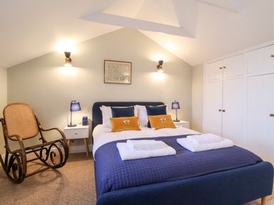 A bedroom with a bed with blue and yellow pillows bedside tables with blue lamps a wooden rocking chair and white wardrobes at Poppy Cottage in Southwold