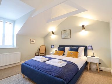A bedroom with a double bed with blue bedding and yellow pillows a wooden chair two bedside tables with blue lamps and framed pictures on walls at Poppy Cottage in Southwold