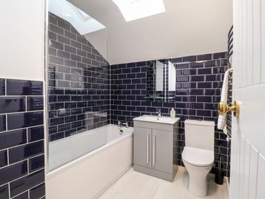 A bathroom with a bathtub sink with cabinet toilet and dark blue tiled walls at Poppy Cottage in Southwold