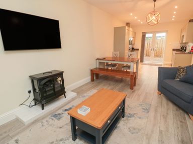 A living room with a wall mounted tv electric stove a wooden coffee table a dining table with bench and chairs a blue armchair and kitchen units at Bamburgh Dunes Bamburgh near Seahouses
