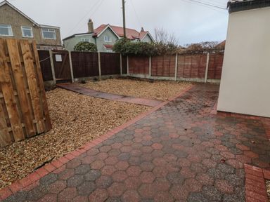 A backyard with a paved walkway, gravel area, wooden fence, and neighboring houses visible in the background