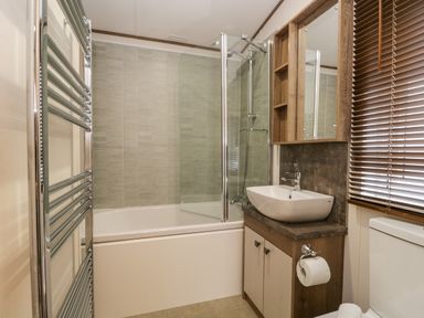 A bathroom with a bathtub and shower, a sink with cabinet, a toilet, a heated towel rail, and a window with blinds at Hudson's Hideaway in South Lakeland Leisure Village