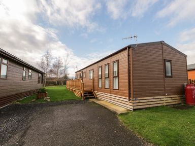 A brown mobile home with stairs and railing on a grassy plot next to a gravel driveway at Hudson's Hideaway in South Lakeland Leisure Village