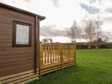 The side of a brown wooden cabin with a window and wooden fence on a grassy area with trees at Hudson's Hideaway in South Lakeland Leisure Village