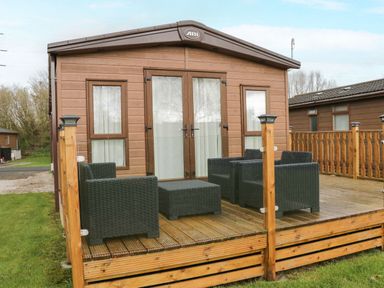 A wooden deck with four black wicker chairs and a small table in front of a brown cabin at Hudson's Hideaway in South Lakeland Leisure Village