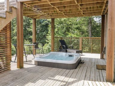 An outdoor deck with a hot tub two chairs a small table and wooden railing surrounded by trees at 4 Faraway Fields in Dobwalls