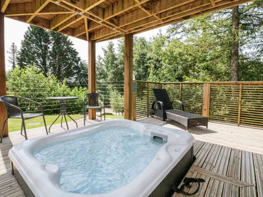 An outdoor covered wooden deck with a hot tub two chairs a small round table and a lounge chair surrounded by trees at 4 Faraway Fields in Dobwalls