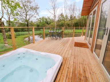 A wooden deck with a hot tub table and chairs overlooking a pond and trees at 21 Meadow Retreat in Dobwalls
