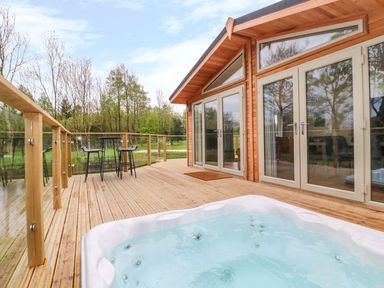 A wooden deck with a hot tub and outdoor seating next to a cabin with glass doors at 21 Meadow Retreat in Dobwalls