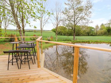 A wooden deck with a table and chairs overlooking a pond and grassy fields at 21 Meadow Retreat in Dobwalls