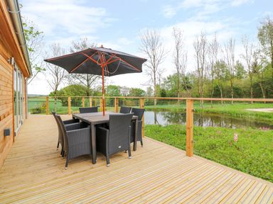 A wooden deck with a dining table set and umbrella overlooking a pond and trees at 21 Meadow Retreat in Dobwalls