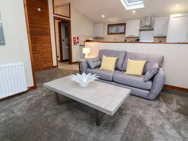 A living room with a gray sofa with yellow cushions a rectangular coffee table and kitchen area in the background at 2 Southern Place in Dobwalls