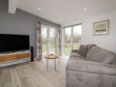 A living room with a gray couch, TV on a wooden stand, coffee table, and large windows at 19 Meadow Retreat in Dobwalls