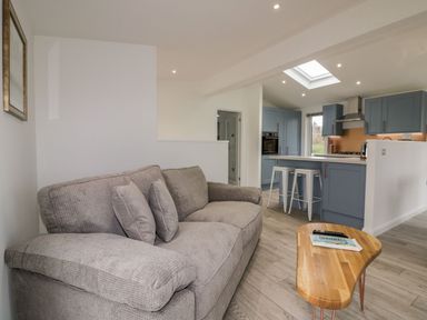 A living area with a gray sofa and wooden table facing a blue kitchen with stools and appliances at 19 Meadow Retreat in Dobwalls