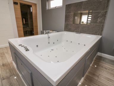 A bathroom with a large white jacuzzi bathtub wooden sauna door and grey tiled wall at 19 Meadow Retreat in Dobwalls