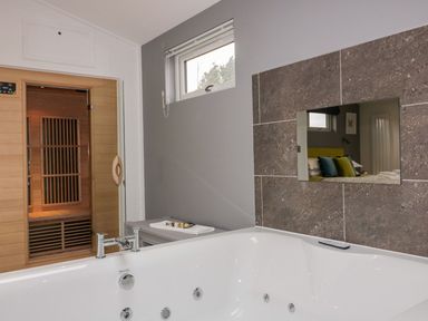 A bathroom with a white bathtub a glass door leading to a wooden sauna and a window on the gray wall at 19 Meadow Retreat in Dobwalls