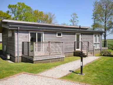 A wooden cabin with a small porch and gravel pathway surrounded by grass and trees at 19 Meadow Retreat in Dobwalls