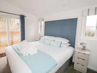 A bedroom with a bed with blue headboard and light blue cushions next to windows and a nightstand with a lamp at 1 Southern Place in Dobwalls