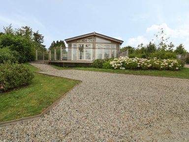 A gravel driveway leading to a small house with glass doors and a wooden deck surrounded by bushes and flowers at 11 Faraway Fields in Dobwalls