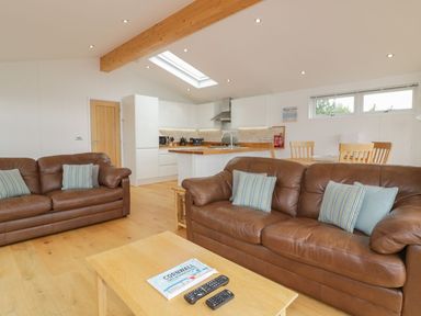 A living room with two brown leather sofas a wooden coffee table with remotes and a kitchen with white cabinets and a wooden breakfast bar at 11 Faraway Fields in Dobwalls