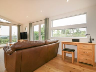 A living room with a brown leather sofa a wooden desk with a lamp and stool and large windows at 11 Faraway Fields in Dobwalls