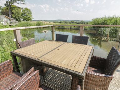 An outdoor deck with a wooden table and six wicker chairs overlooking a pond at 15 Faraway Fields in Dobwalls