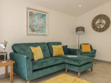 A living room with a green sectional sofa with yellow pillows a yellow rug a small side table with a lamp an armchair with a yellow pillow a floor lamp a clock and a framed abstract painting on the wall at 18 Meadow Retreat in Dobwalls