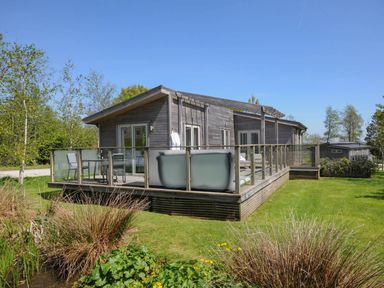 A wooden cabin with a glass railing deck and outdoor seating on a lawn with trees and shrubs at 11 Meadow Retreat in Dobwalls