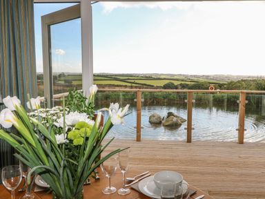 A dining table with flowers cups and plates next to an open door leading to a wooden deck and a pond with rocks and countryside view at 14 Faraway Fields in Dobwalls