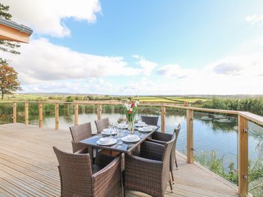 An outdoor dining table with six chairs set on a wooden deck overlooking a pond and countryside at 14 Faraway Fields in Dobwalls