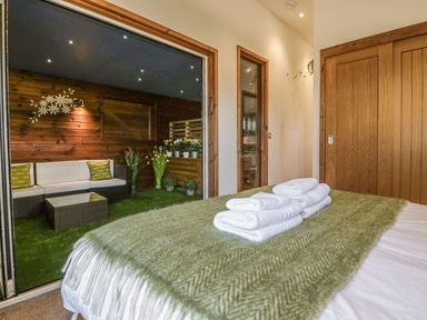 A bedroom with a bed covered by a green blanket and folded white towels near a sliding glass door leading to a seating area with white sofas and potted plants at 9 Meadow Retreat in Dobwalls