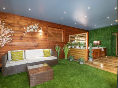 An indoor room with artificial grass flooring a wicker sofa with cushions a wicker table wooden walls and a wooden deck with a spa bath and various potted plants at 9 Meadow Retreat in Dobwalls