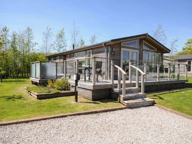 A wooden cabin with a glass railing deck and steps on a grassy lawn at 7 Meadow Retreat in Dobwalls