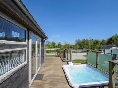 An outdoor wooden deck with a hot tub next to a building and glass railing at 5 Meadow Retreat in Dobwalls