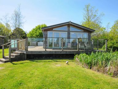 A wooden cabin with glass railing and outdoor seating on a deck overlooking grass and reeds at 5 Meadow Retreat in Dobwalls