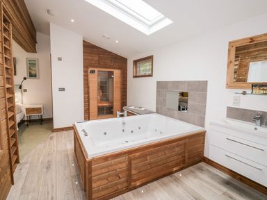 A bathroom with a large wooden-framed jacuzzi tub a wooden sauna bench a wall-mounted sink with mirror and a door leading to a bedroom at 2 Meadow Retreat in Dobwalls