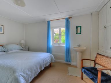 A bedroom with a bed a bedside table and lamp a window with blue curtains a washbasin and a chair at 30 Sherborne Lane Lyme Regis