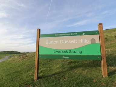 A sign for Burton Dassett Hills with grass and a road under a blue sky