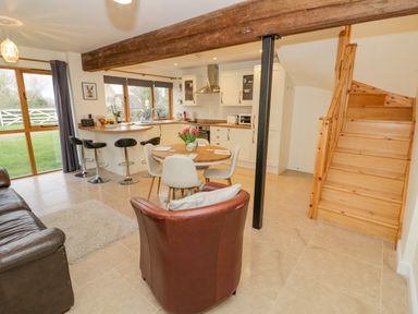 An open plan room with a kitchen, dining table, sofa, armchair, bar stools, and wooden staircase at The Granary in Little Dasset near Gaydon