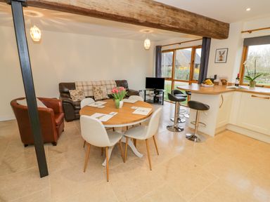 An open plan living area with a round dining table and chairs a brown sofa a leather armchair bar stools at a kitchen counter and large windows showing a garden at The Granary Little Dasset near Gaydon