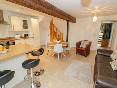 An open plan kitchen and living area with a round dining table and chairs a leather sofa an armchair bar stools and wooden stair at The Granary in Little Dasset near Gaydon