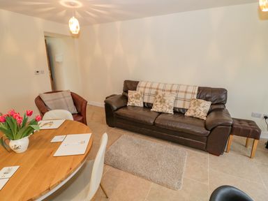 A living room with a brown leather sofa with cushions and a beige throw a wooden dining table with chairs and a vase of pink tulips at The Granary Little Dasset near Gaydon