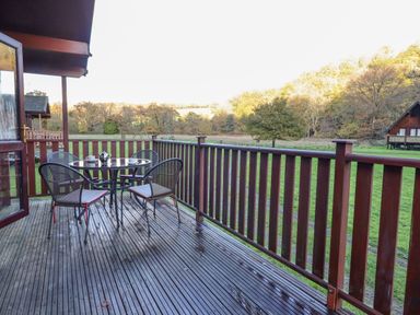 An outdoor wooden deck with a round glass table and four chairs overlooking a grassy field and trees at Dartmoor Retreat Lodge Clifford Bridge near Moretonhampstead